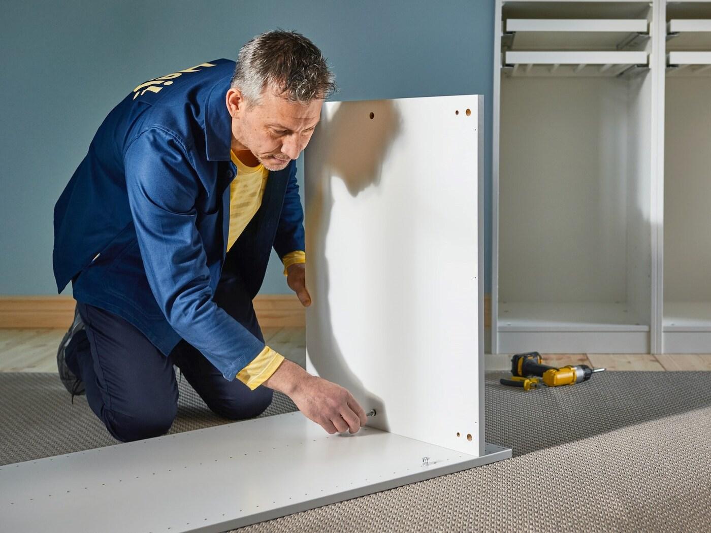 Un montatore con abbigliamento da lavoro IKEA blu e giallo è inginocchiato sul pavimento e monta la struttura di un armadio.