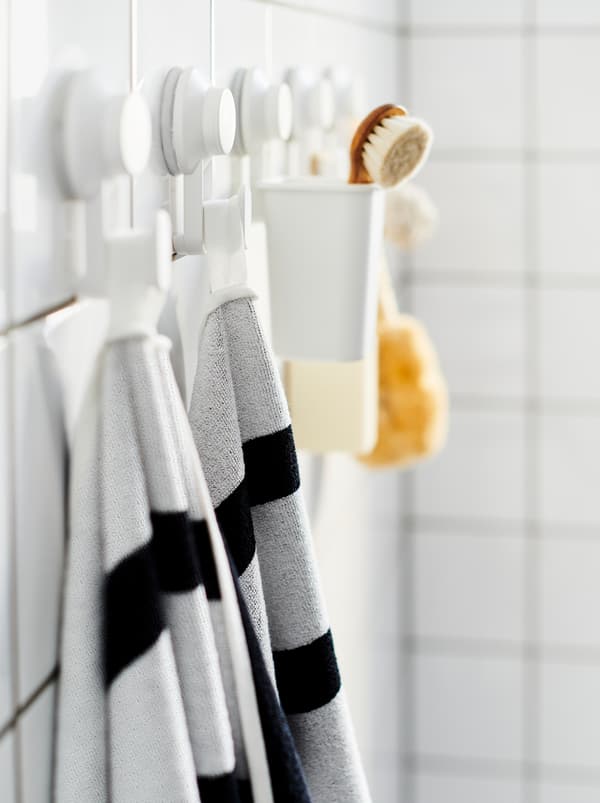 Un mur de salle de bains aux carreaux blancs avec une rangée de crochets TISKEN où sont accrochés des serviettes, une brosse et d’autres accessoires de salle de bains.
