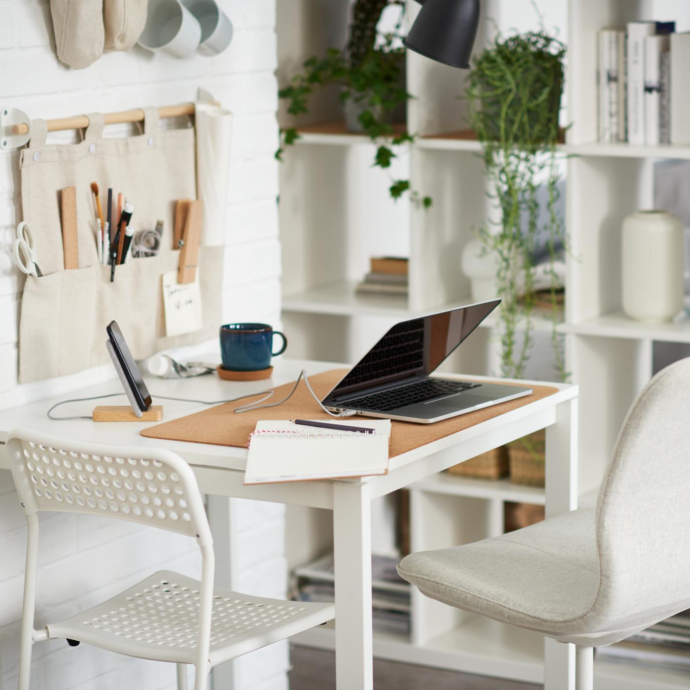 Un ordinateur portable sur une table blanche avec une chaise blanche et une chaise beige, une étagère blanche accueillant des livres, des plantes et des rangements.
