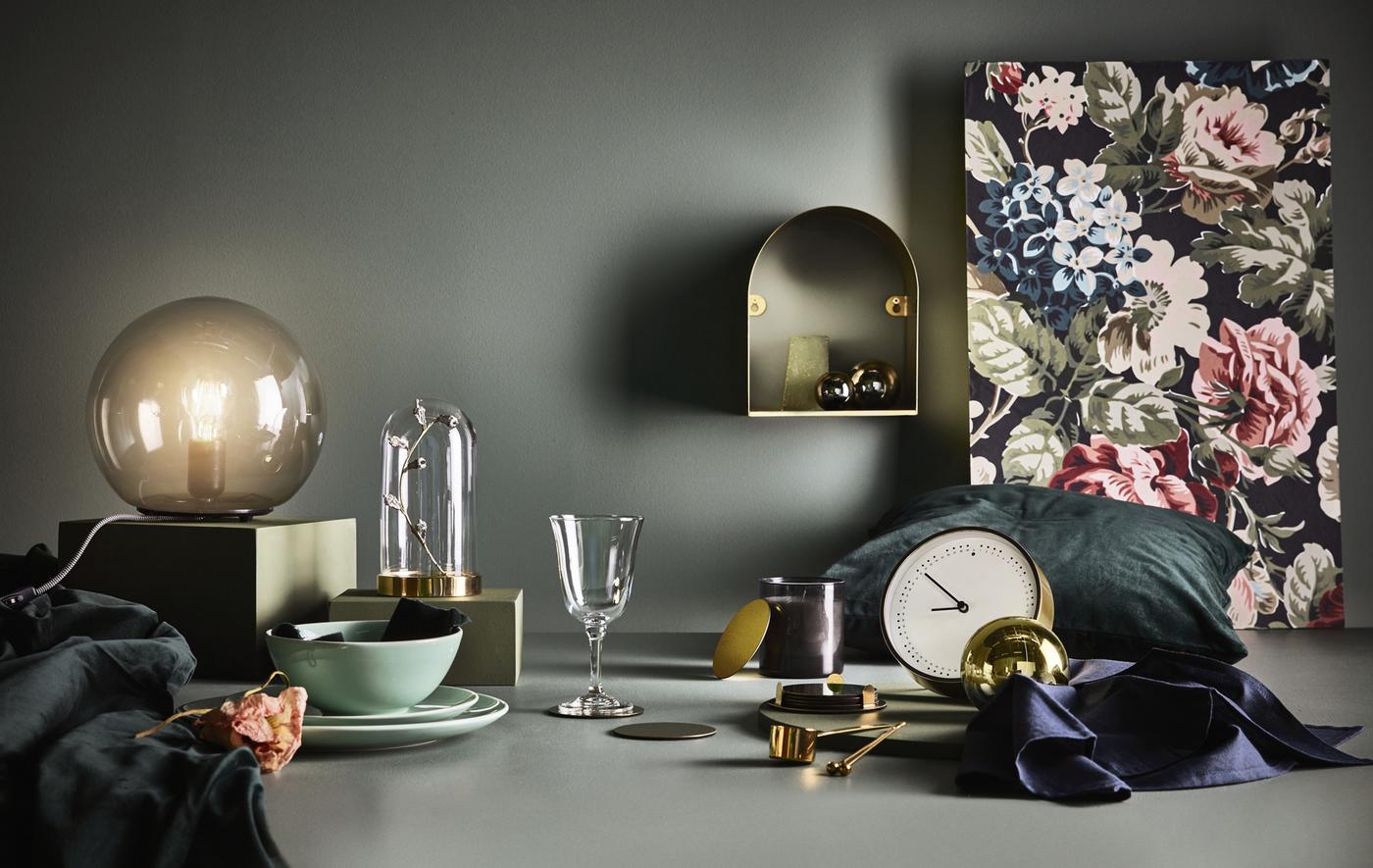 Un orologio, una lampada da tavolo, una campana di vetro e tanti altri oggetti disposti davanti a uno sfondo scuro - IKEA
