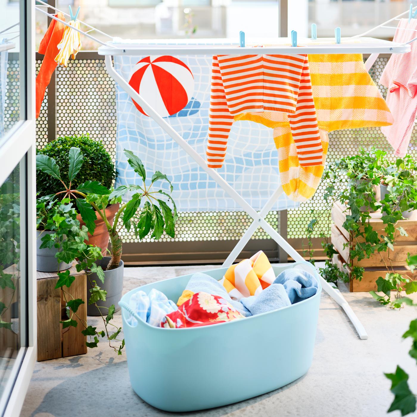 Un panier à linge souple TORKIS bleu rempli de linge et un séchoir blanc sur un balcon ensoleillé avec des plantes vertes.