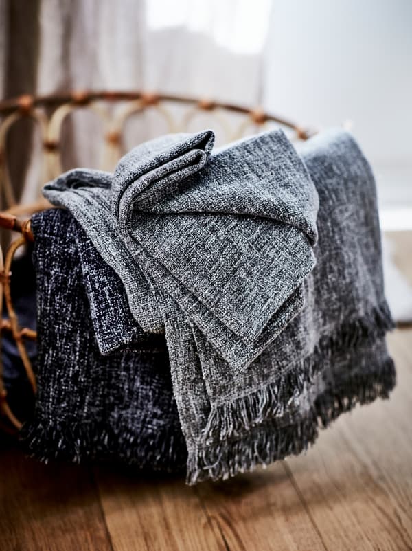 Un panier en rotin tressé brun clair sur un sol en bois contenant des plaids RÖNNBÄRSMAL pliés et débordant sur le côté.