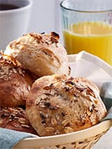 Un panier rempli de petits pains fermentés à froid avec du muesli et des flocons de sel. À l'arrière, il y a un verre de jus d'orange.