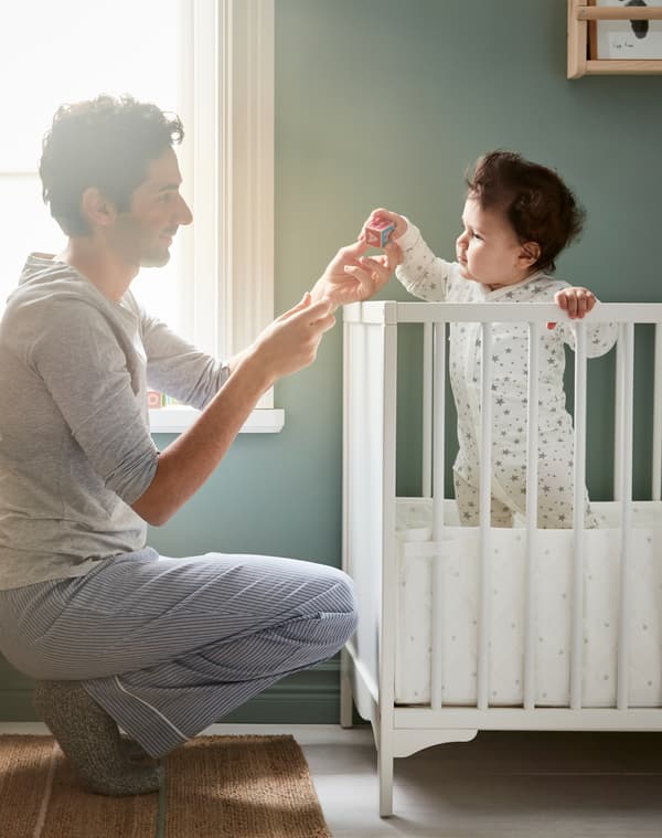Un papà gioca con un peluche insieme al suo bambino, in piedi in un lettino bianco SUNDVIK davanti a una finestra illuminata dal sole - IKEA