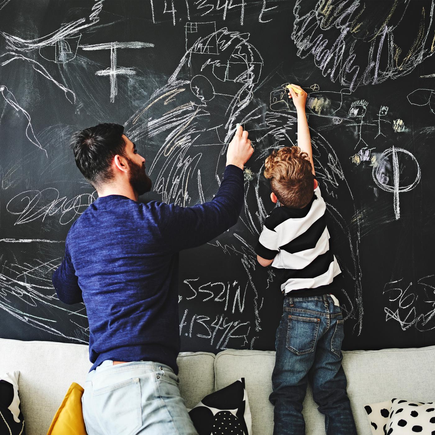 Un parent et un enfant sont à genoux et debout sur un canapé. Ils dessinent sur un mur recouvert d’une peinture à tableau.