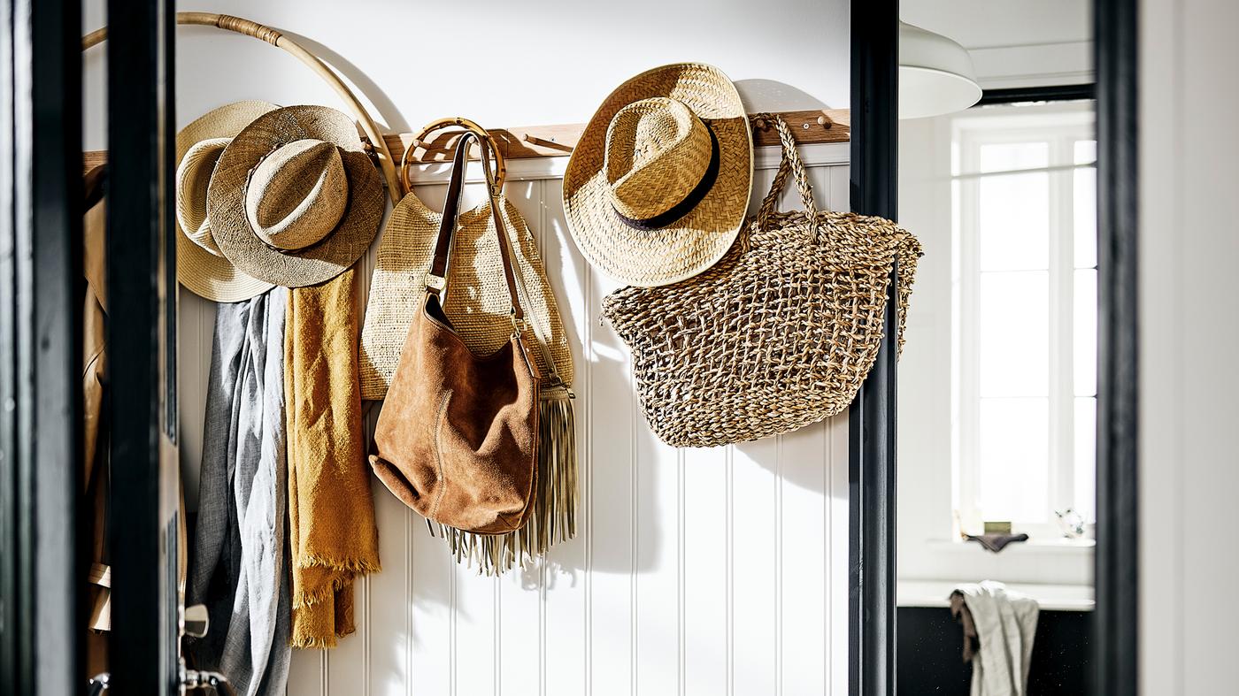 Un pasillo blanco con un perchero con seis pomos que sostiene bolsos, sombreros y bufandas en colores naturales, y un espejo TOFTBYN al lado.