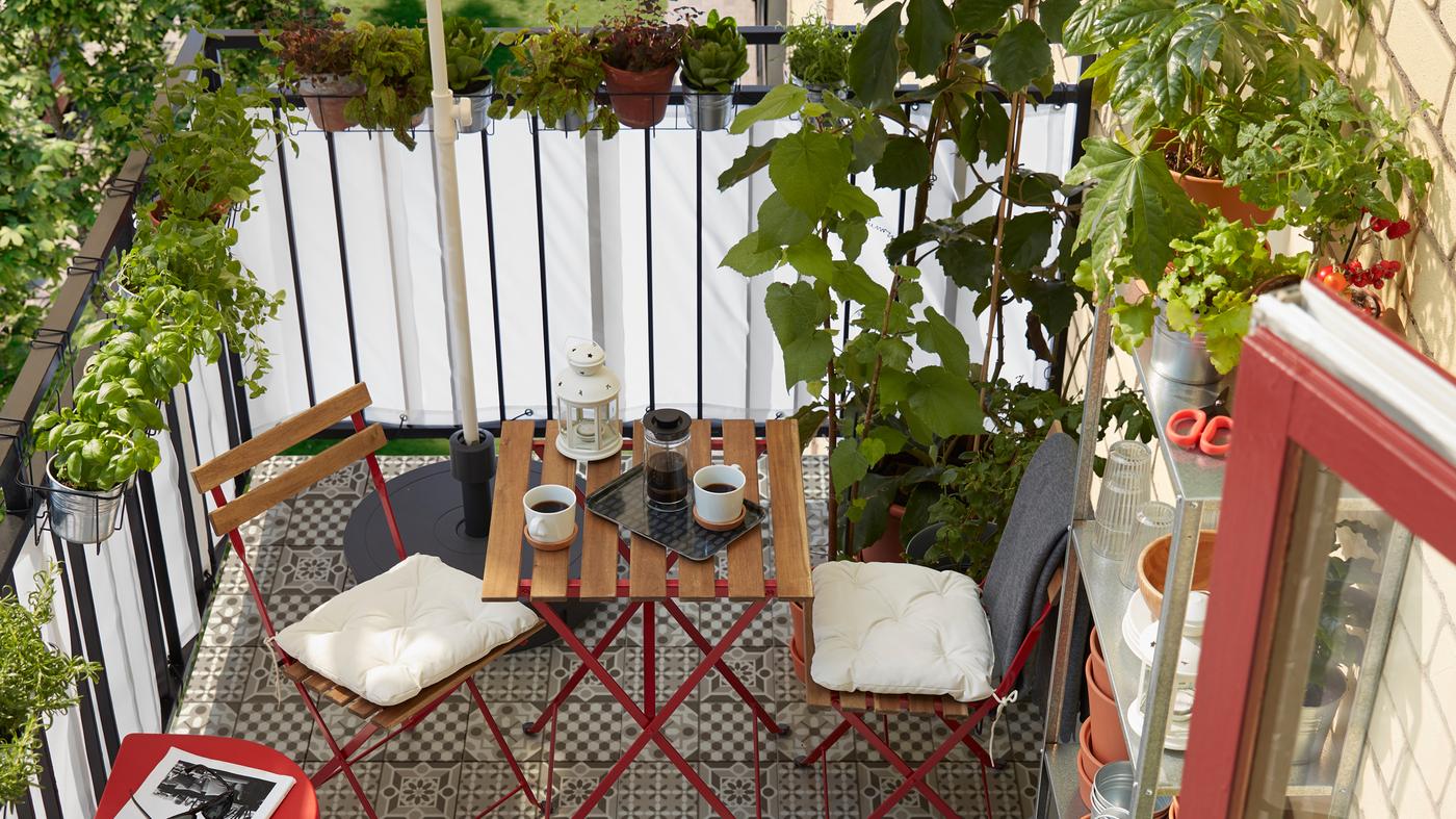 Un pequeño balcón con muchas hierbas y plantas, una estantería con macetas, un parasol, dos sillas y una mesa.