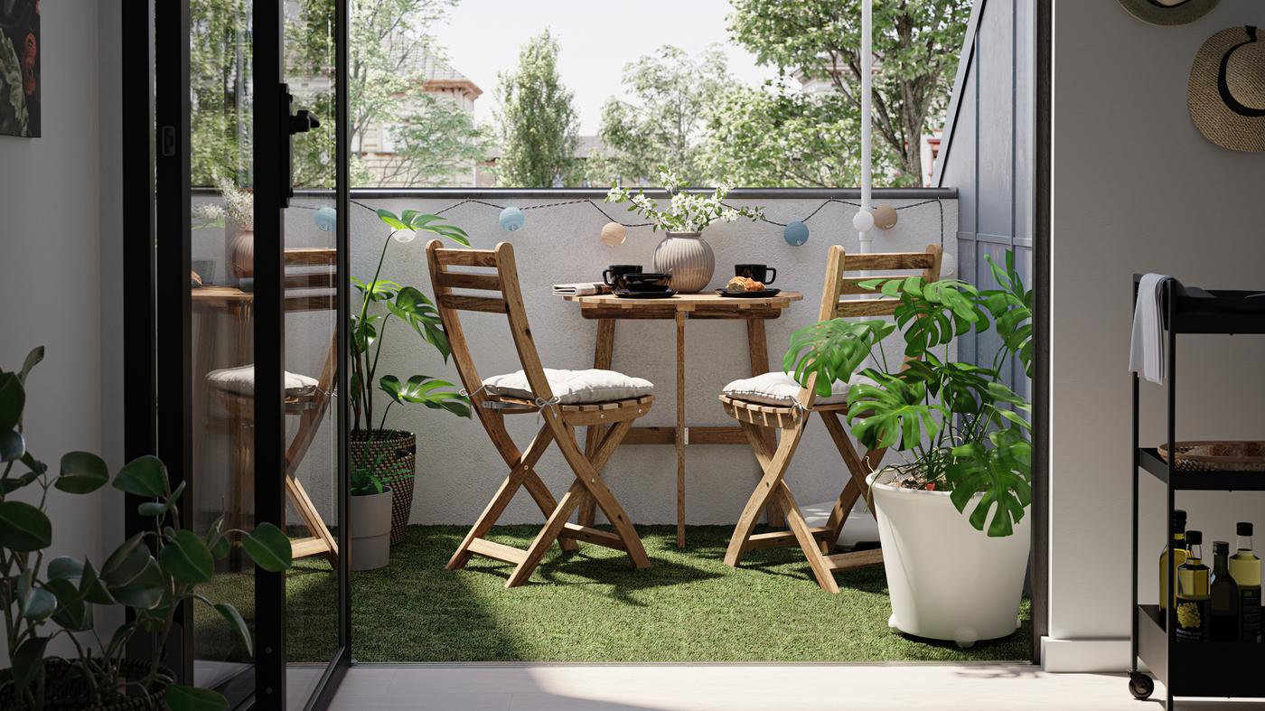 Un petit balcon avec une table et des chaises en bois, un revêtement de sol imitation gazon et un Monstera dans un cache-pot blanc.