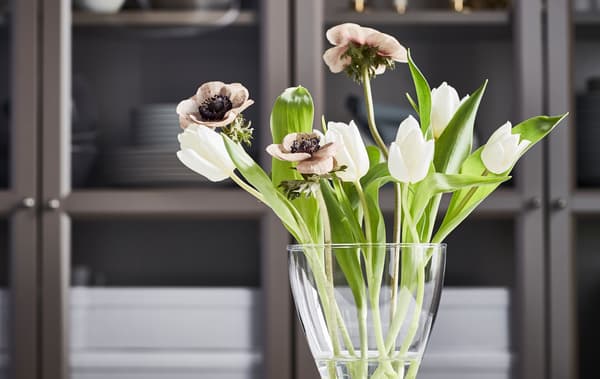 Un petit bouquet de tulipes blanches, de coquelicots brunâtres et de feuilles vertes fraîches dans un vase VASEN arrondi, devant une armoire.