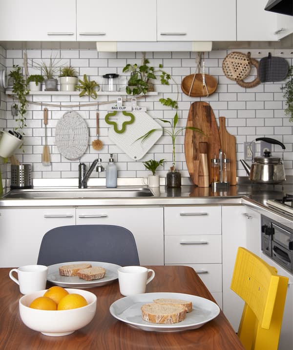Un petit-déjeuner simple posé sur une table en bois dans une petite cuisine blanche avec des plantes vertes et des ustensiles accrochés aux murs.