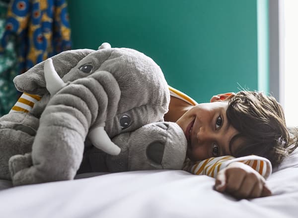 Un petit garçon heureux couché sur un lit dans une chambre à coucher vert foncé avec une peluche éléphant grise JÄTTESTOR.