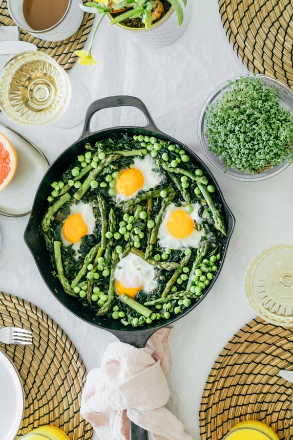 Un piatto vivace preparato in padella con asparagi, piselli e uova, servito su un tavolo bianco con tovagliette intrecciate, circondato da altri piatti e da un'insalata verde, evoca un brunch fresco e accogliente.