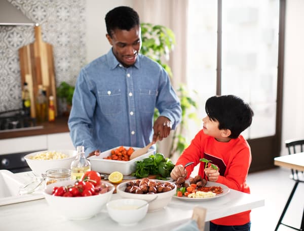 Un plan de travail de cuisine chargé de saladiers de carottes cuites, de pâtes, de boulettes végétales et de tomates. Un adulte sert à manger à un enfant.
