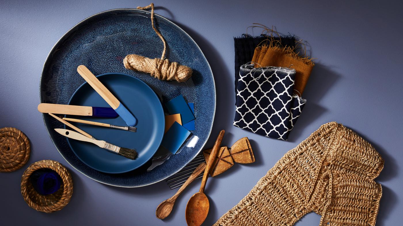 Un plat ERTAPPAD bleu tenant un rouleau de ficelle et des pinceaux, à côté d'échantillons de tissu, de cuillères en bois et d'objets tissés.