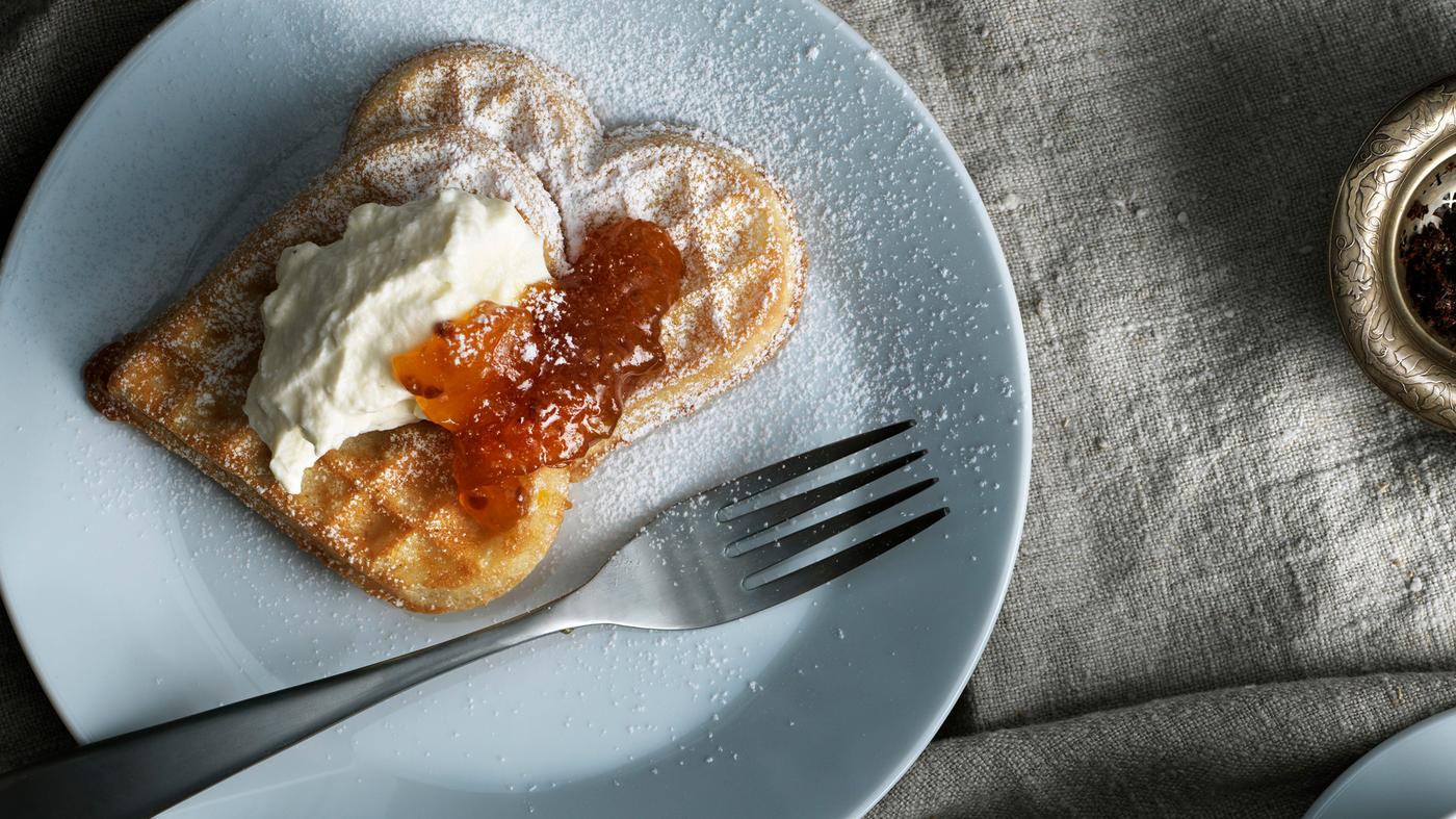 Un plato blanco con un tenedor y dos waffles en forma de corazón, con crema y mermelada de naranja. El azúcar en polvo se espolvorea encima.