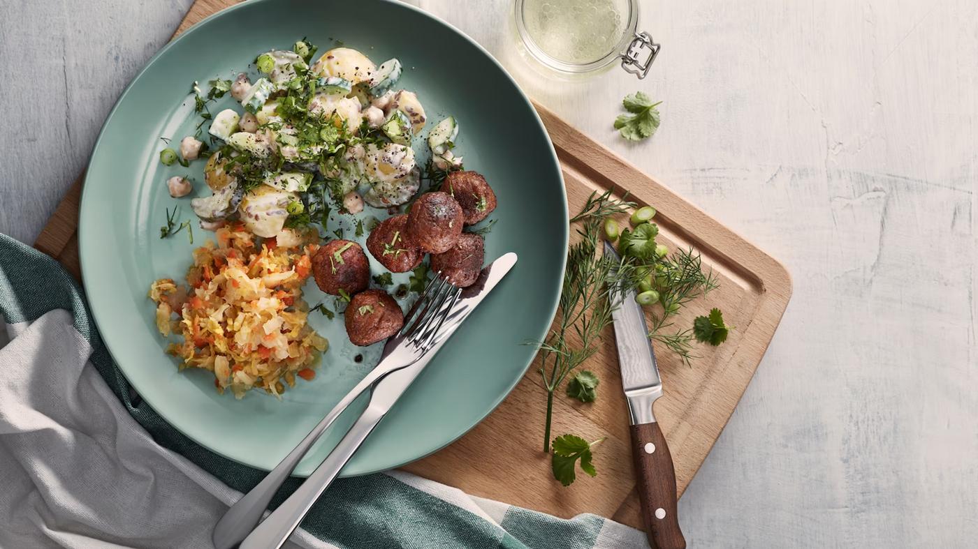 Un plato turquesa con albondigas vegetales, ensalada de papas y kimchi, todo sobre una tabla para cortar con un cuchillo, hierbas y paños de cocina.