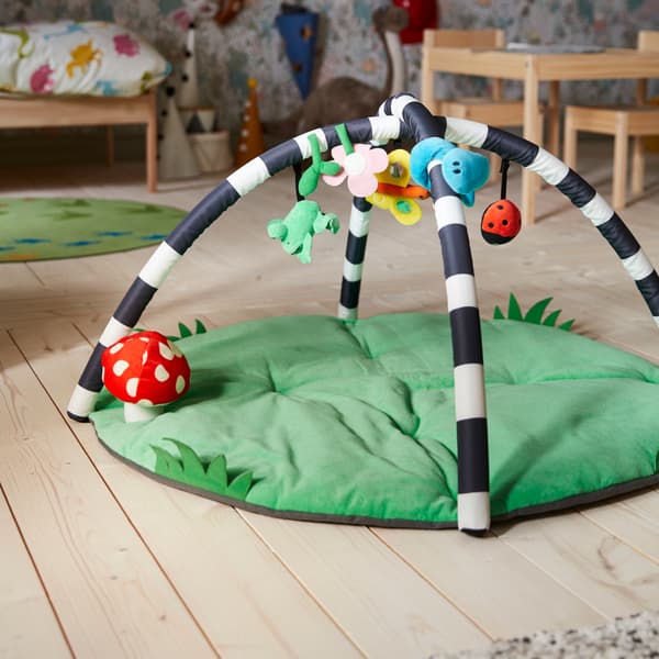 Un portique bébé KLAPPA posé sur un sol en bois clair. Derrière lui, un lit bébé, une table et des chaises pour enfant, et des jouets.