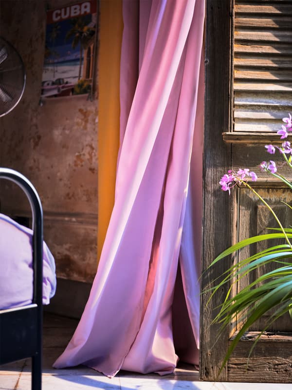 Un rideau purificateur d’air, mauve clair, flottant au vent à côté d’une persienne en bois.