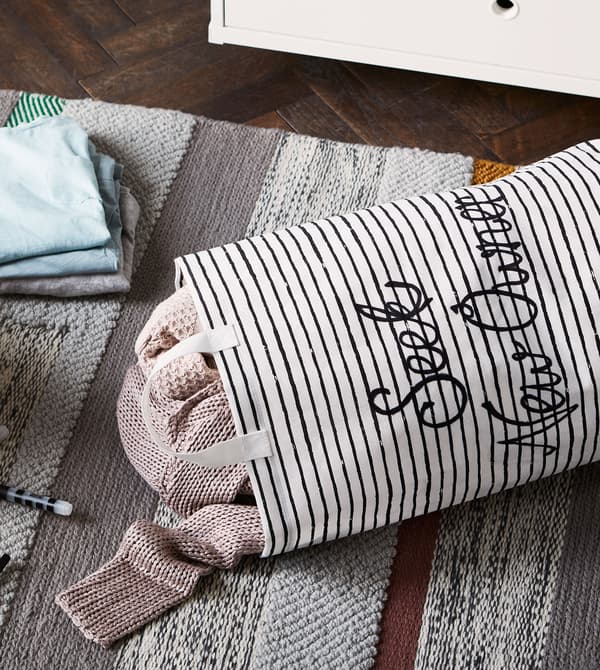 Un sac à lessive KLUNKA rayé rempli et une pile de vêtements sur un tapis gris.