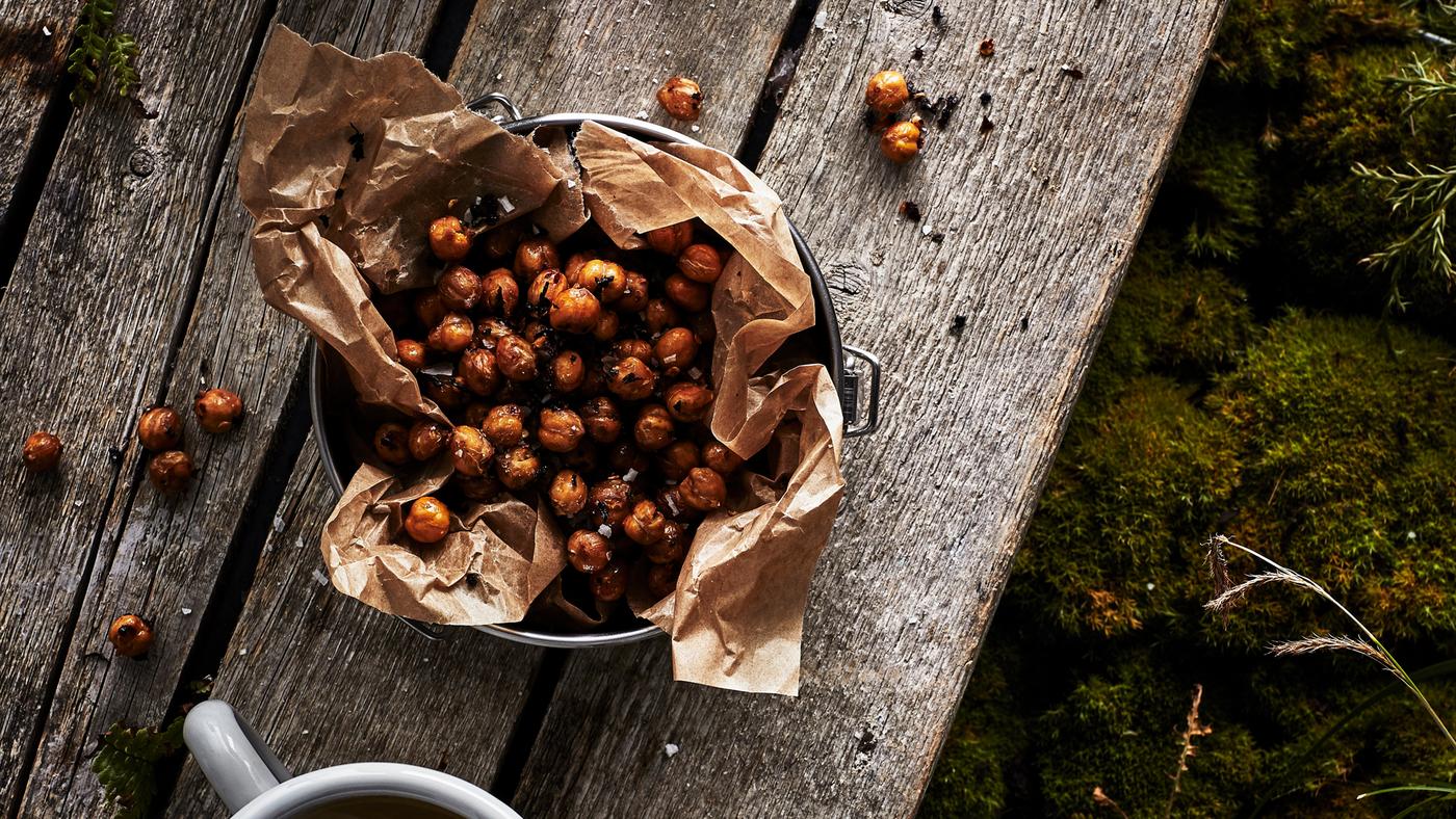 Un saladier garni de papier placé sur une table en bois rustique et rempli de pois chiches grillés au chaï et aux piments. Une tasse se trouve à proximité.