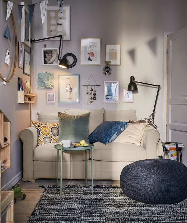 Un séjour avec un canapé deux places, un pouf bleu foncé, une petite table plateau grise et des lampes noires de chaque côté.