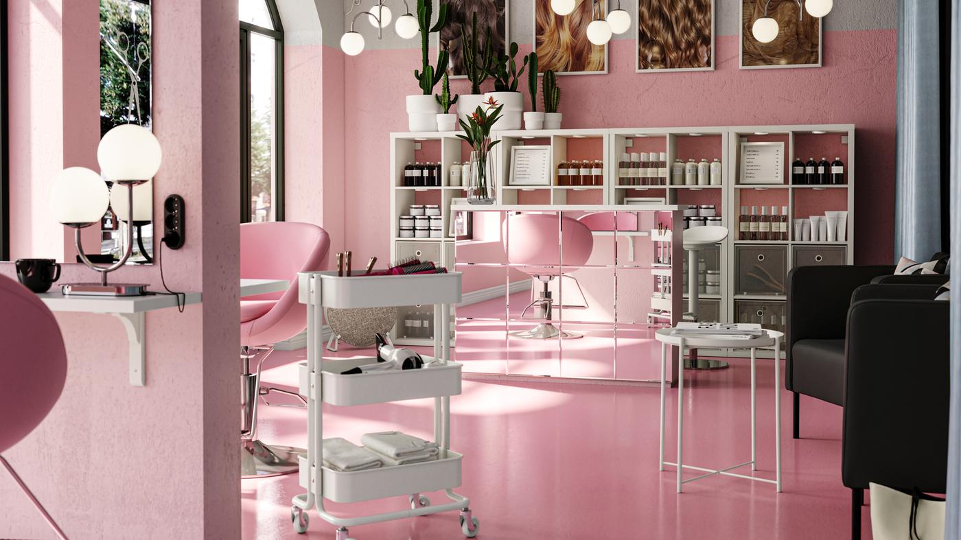 Un spacieux salon de coiffure rose avec des étagères KALLAX derrière un bureau de réception à miroirs. Une desserte RÅSKOG se trouve près d’un poste de travail.
