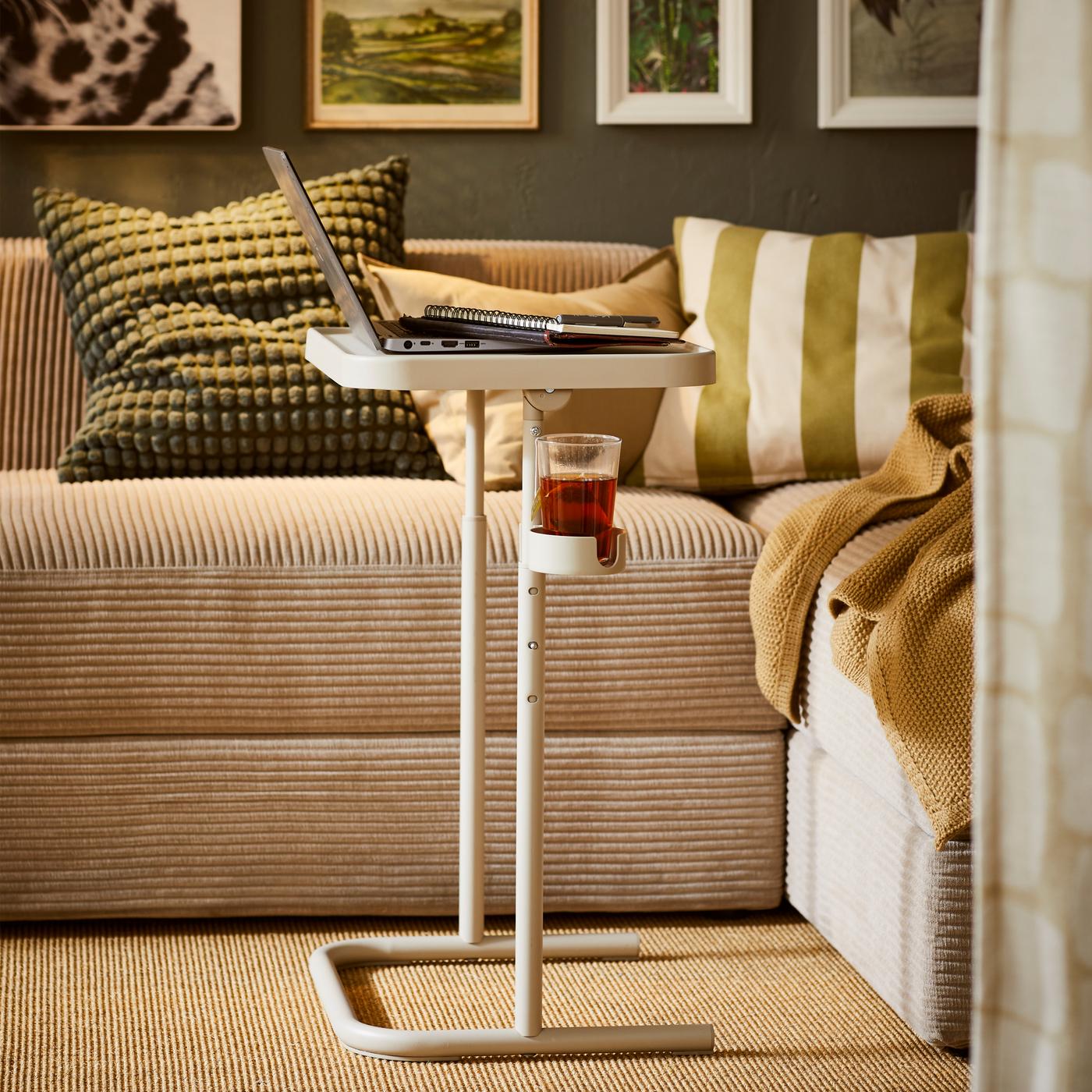 Un support pour ordinateur portable BJÖRKÅSEN beige situé près d’un canapé JÄTTEBO gris-beige agrémenté de textiles. Une rangée d’œuvres artistiques encadrées au-dessus d’un canapé.