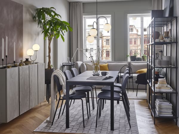 Un tavolo nero/marrone scuro e sedie grigie su un tappeto a motivi geometrici. Sopra, una lampada a sospensione cromata a 3 bracci - IKEA