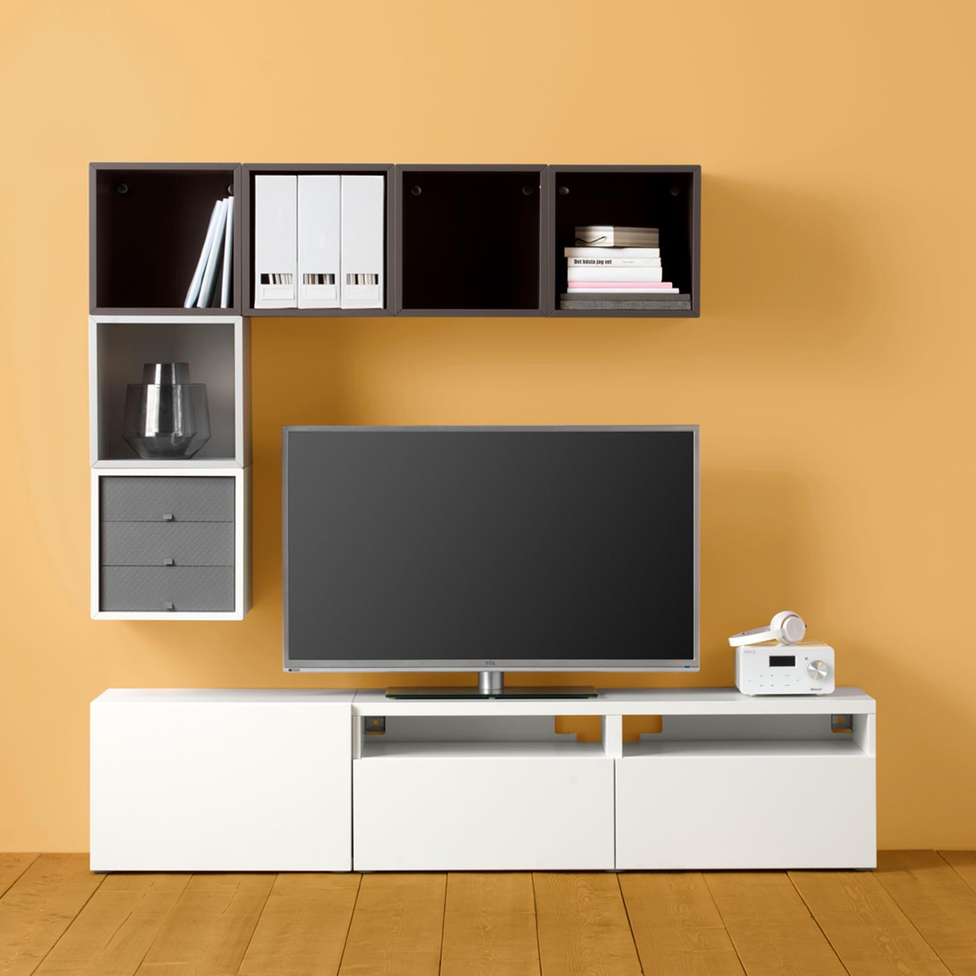 Un téléviseur entouré par trois côtés d’un agencement de rangements BESTÅ et EKET géométrique, mais asymétrique, contre un mur jaune.