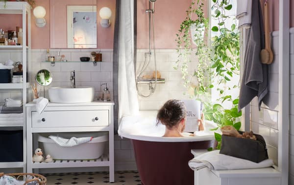 Un uomo nella vasca in un bagno con pareti rosa pallido, piastrelle e lavabo bianchi, piante - IKEA