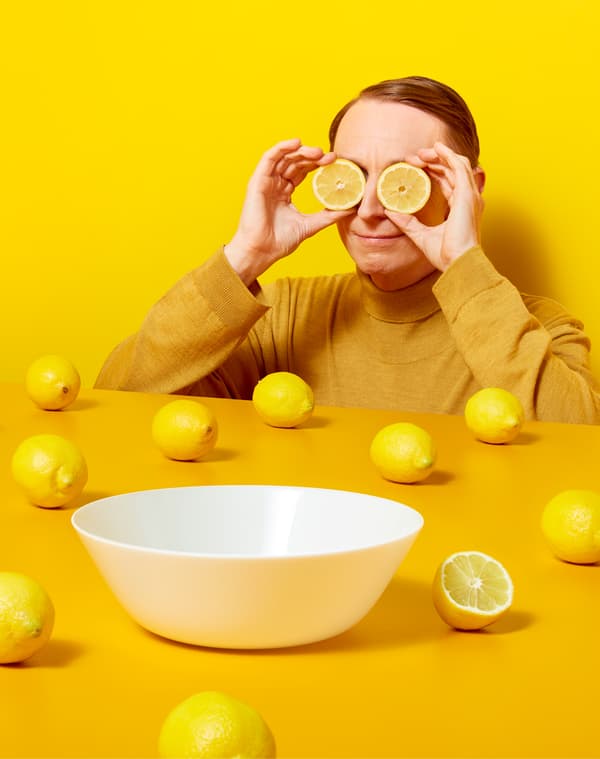 Un uomo vestito di giallo tiene due fette di limone davanti agli occhi, vicino a un tavolo giallo con sopra tanti limoni e una ciotola bianca - IKEA