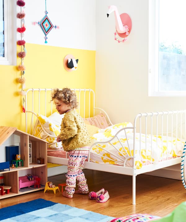 Una bambina in una stanza con una parete gialla, una struttura letto bianca, copripiumino e federe bianche e gialle e una casa delle bambole in legno - IKEA