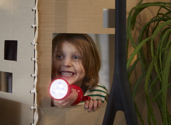 Una bambina sorridente che gioca a nascondino tenendo in mano una torcia manuale LJUSA rossa – IKEA