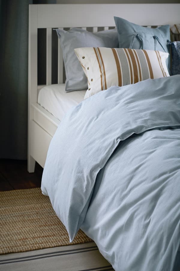 Una cama con un velador, ambos blancos. Se ven cojines de telas con colroes claros, como celeste y blanco principalmente. La cómoda es HEMNES de IKEA y el cajón organizador es STUK.