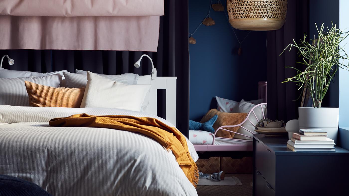 Una cama extensible junto a una cama blanca con ropa de cama beige/blanca, un cojín beige oscuro, cortinas rosas y una lámpara de techo hecha de bambú.