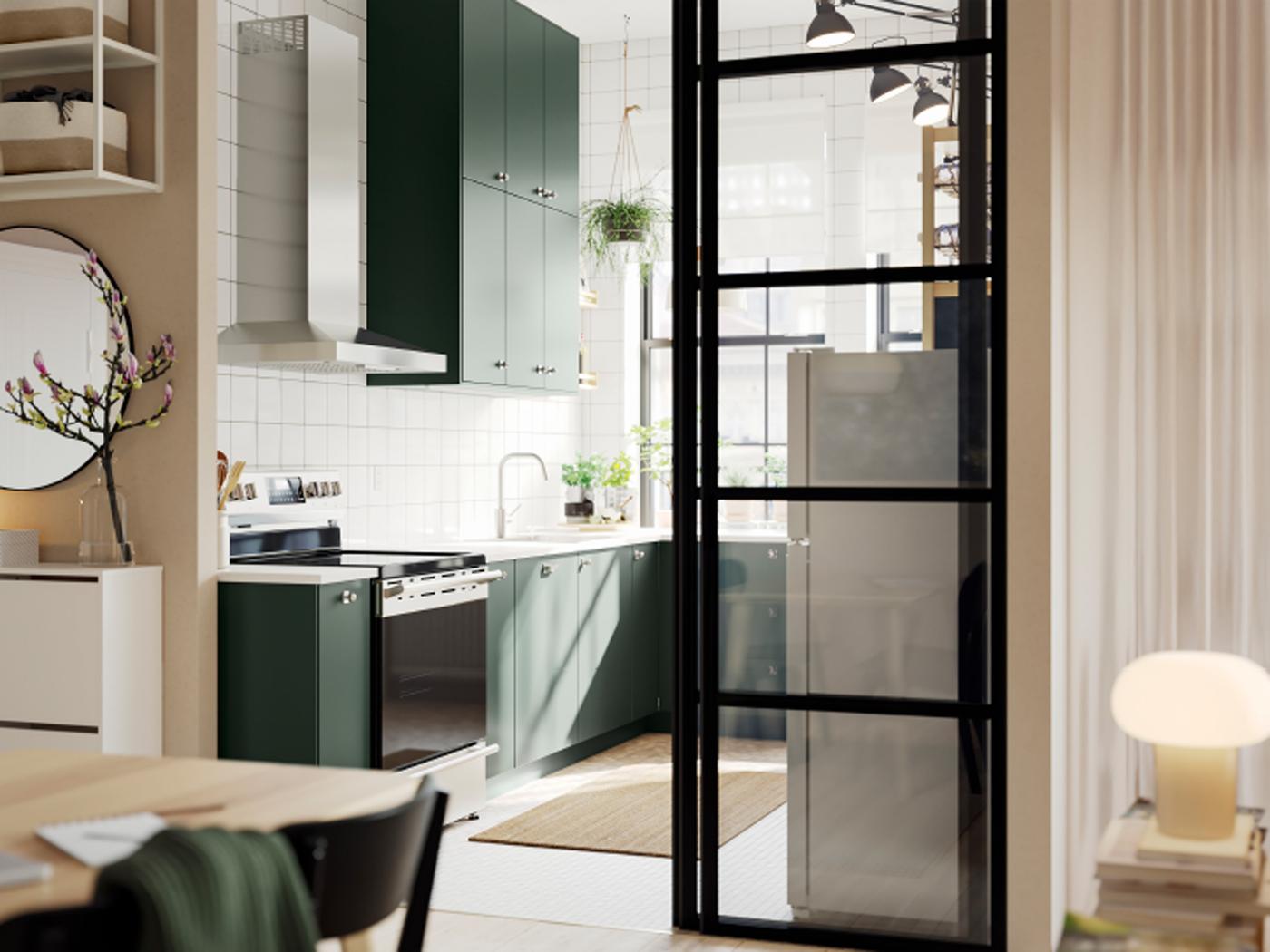 Una cocina abierta con frentes de cocina HAVSTORP en verde oscuro y gabinetes de pared hasta el techo junto a una sala luminosa.