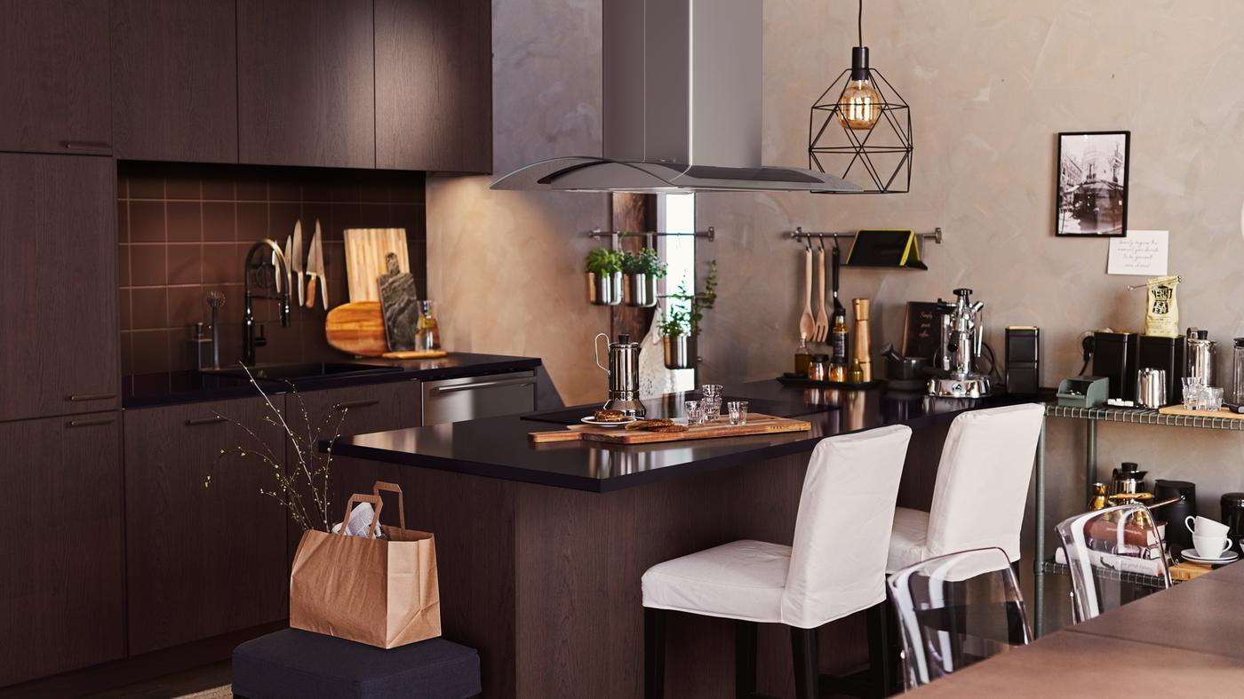 Una cocina color café oscuro con gabinetes SEKTION y una isla. Bancos altos blancos en la isla, y una lámpara BRUNSTA colgada en lo alto.
