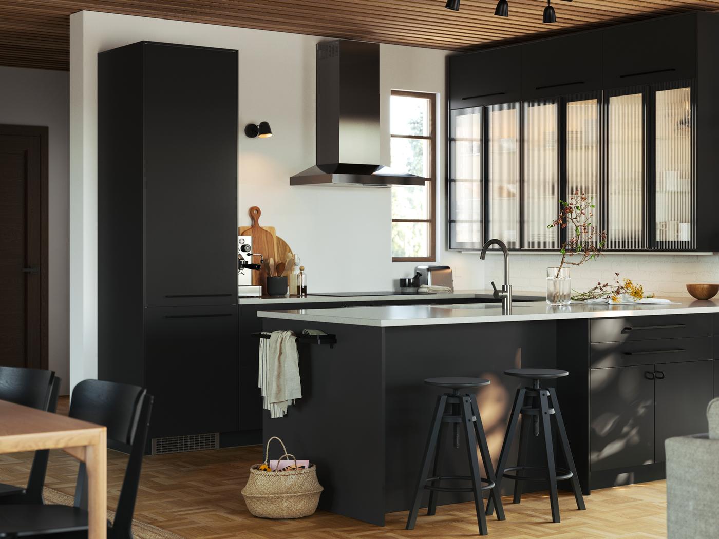 Una cocina con paneles de FÖRBÄTTRA, gabinetes HEJSTA con puertas de cristal en vidrio carbón/redondeado y bancos altos DALFRED en negro.