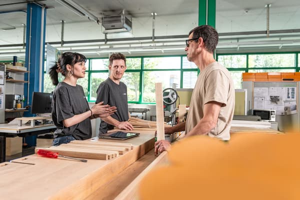 Una collaboratrice e due collaboratori dell’impresa sociale BAND sono in piedi davanti a un banco da lavoro e si consultano. 