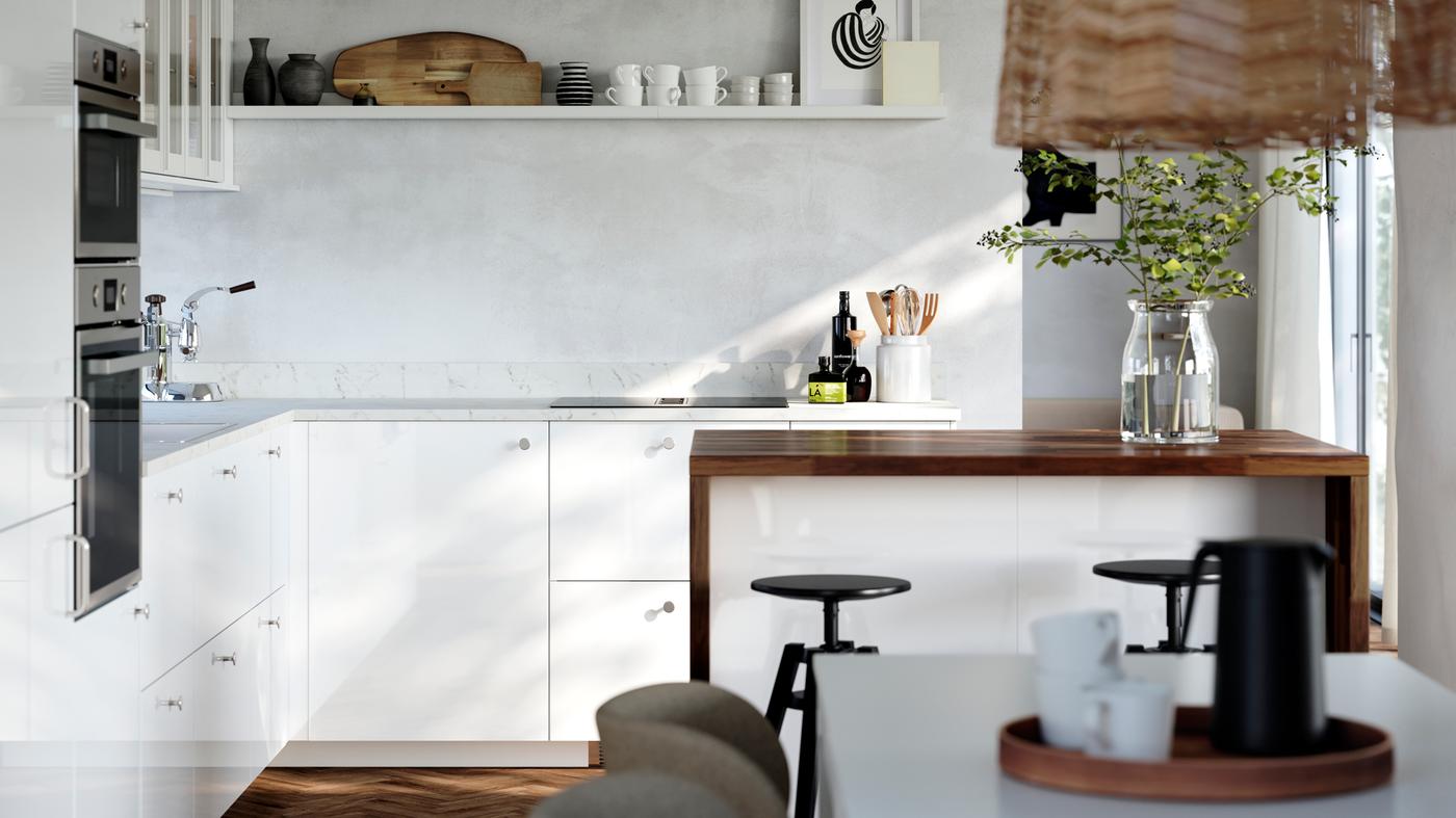 Una cucina con frontali RINGHULT bianco lucido, ante a vetro HEJSTA, una mensola espositiva fissata alla parete e un'isola.