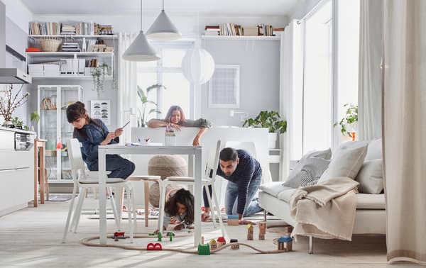 Una familia en su living comedor interactuando. Niños jugando en el suelo, rodeados de mesas y sillas blancas. Lámparas colgando desde el techo blancas con formas de conos y esféras. Sofá estilo futón beige con cojines grises claros.