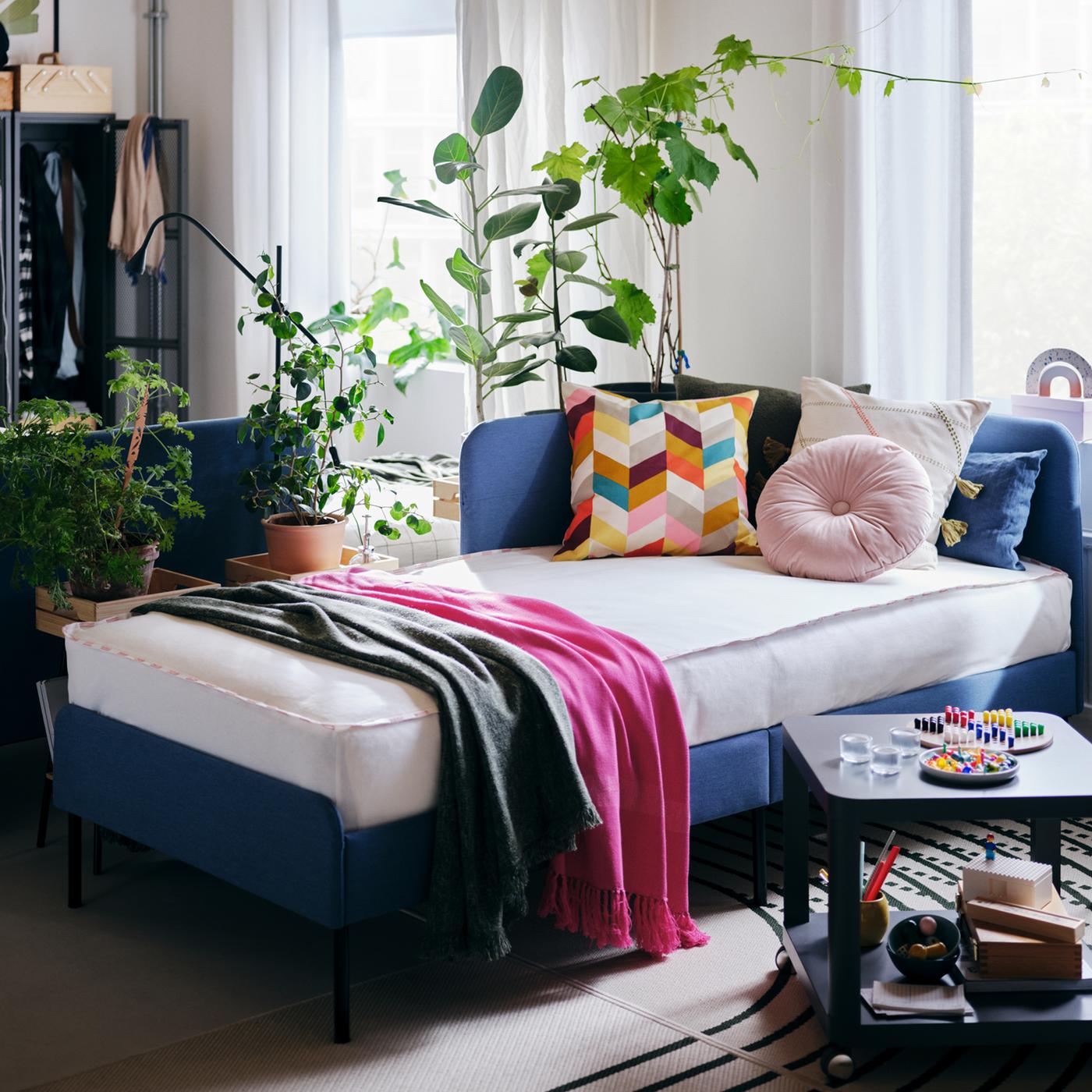 Una habitación con una cama BLÅKULLEN con cojines multicolores, una mesa de centro, ventanas con cortinas y plantas verdes.