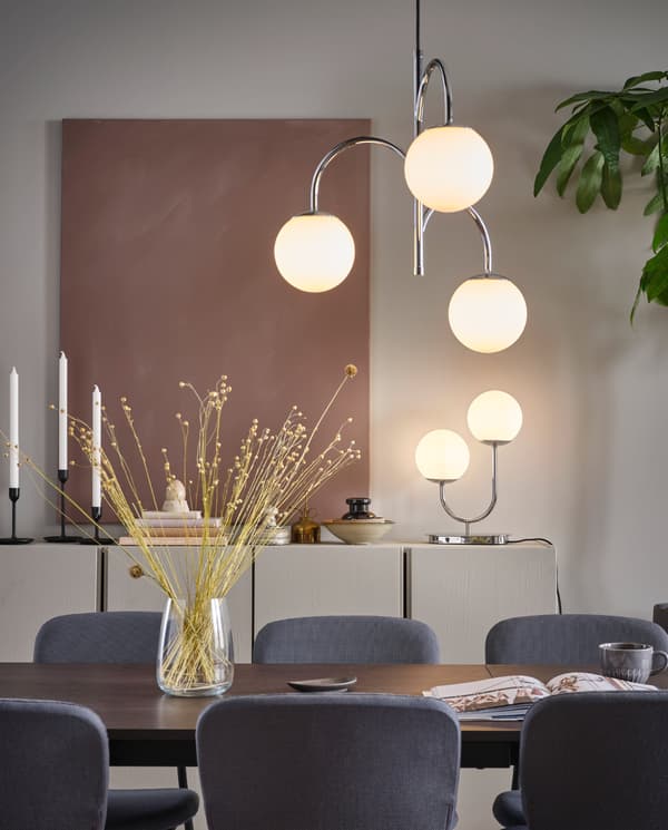 Una lampada a sospensione cromata a 3 bracci SIMRISHAMN appesa sopra un tavolo da pranzo e una lampada da tavolo SIMRISHAMN appoggiata su un mobile - IKEA