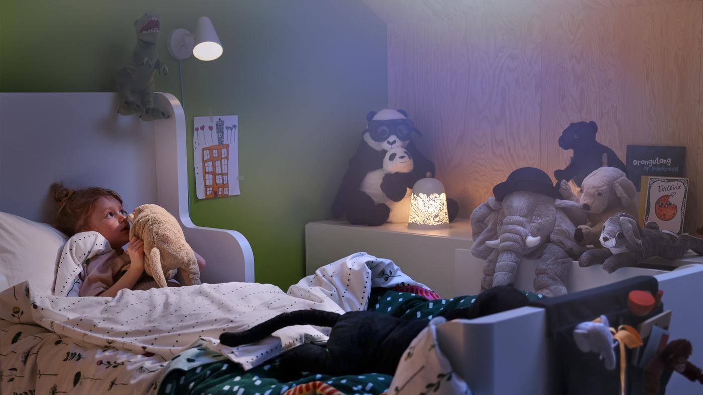 Una lampada da parete FUBBLA e una lampada da tavolo SOLSKUR illuminano la cameretta di una bambina sdraiata a letto che sussurra all’orecchio del suo peluche preferito - IKEA