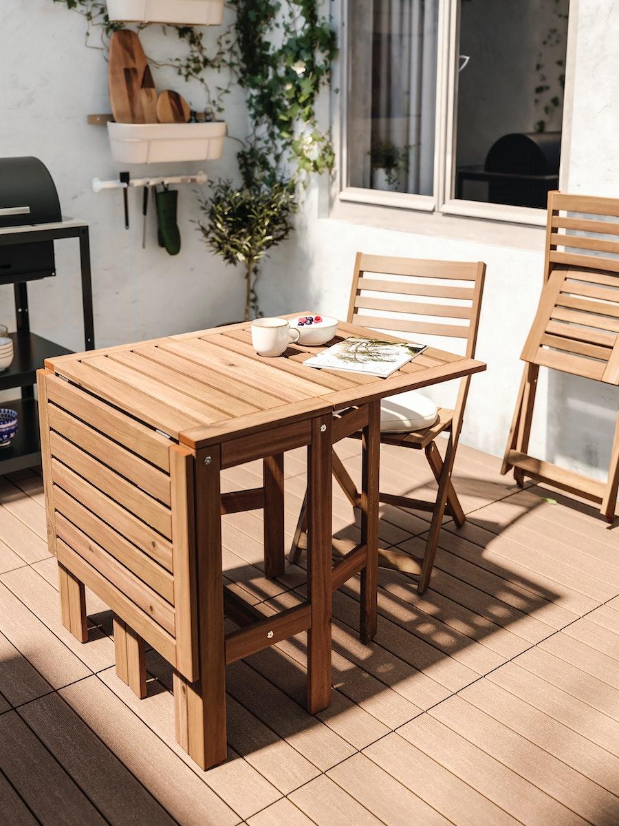 Una mesa de madera ÄPPLARÖ con un extremo plegado, frente a una silla plegable ÄPPLARÖ, en una terraza con piso de madera y muros blancos. Frente a la silla y sobre la mesa hay una revista, un tazón GLADELIG y un bowl de desayuno. 