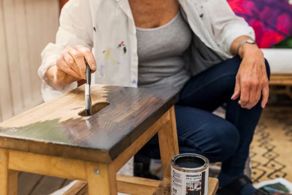Una mujer con polera gris y camisa blanca con manchas de pintura. Está pintando negro un piso de madera.