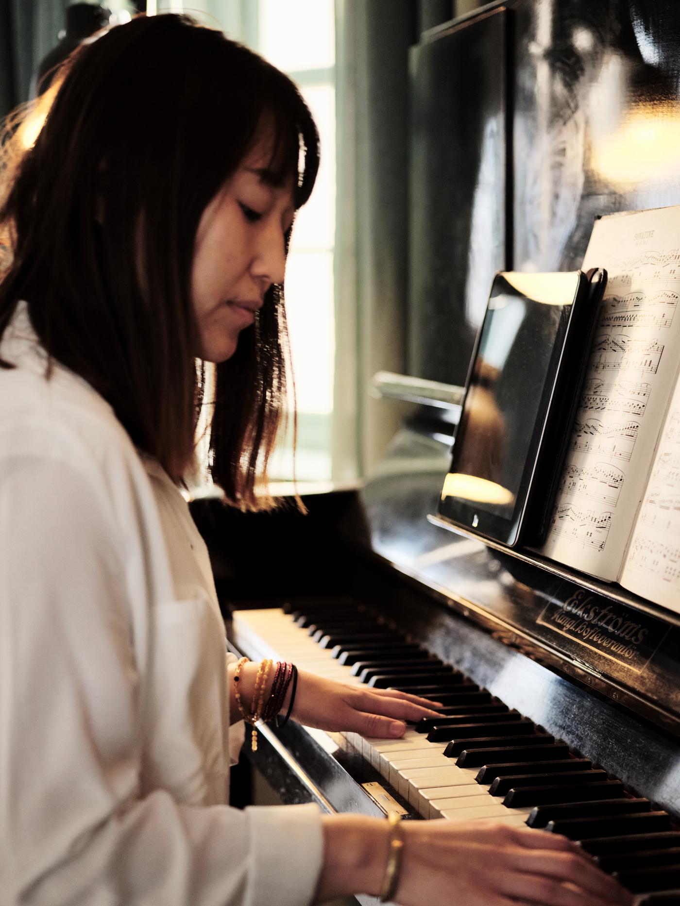 Una mujer de cabello largo y oscuro sentada junto a un piano, con las manos en las teclas.