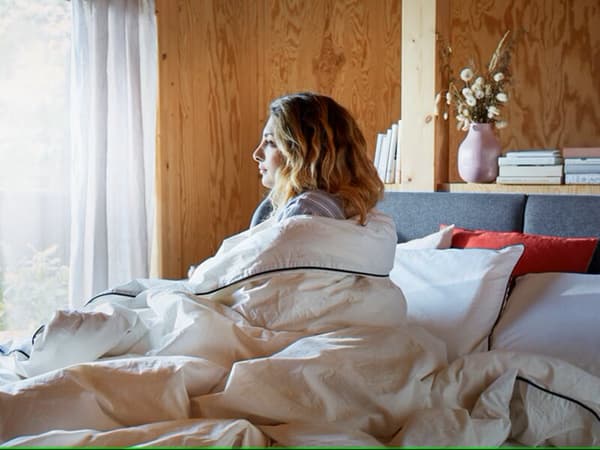 Una mujer envuelta en un plumón IKEA sentada sobre una cama mirando a la ventana.