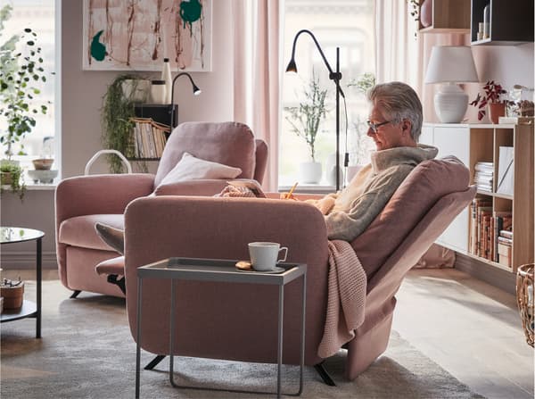 Una mujer sentada cómodamente en el sillón reclinable EKOLSUND con funda GUNNARED café claro y rosa junto a una mesa de centro.