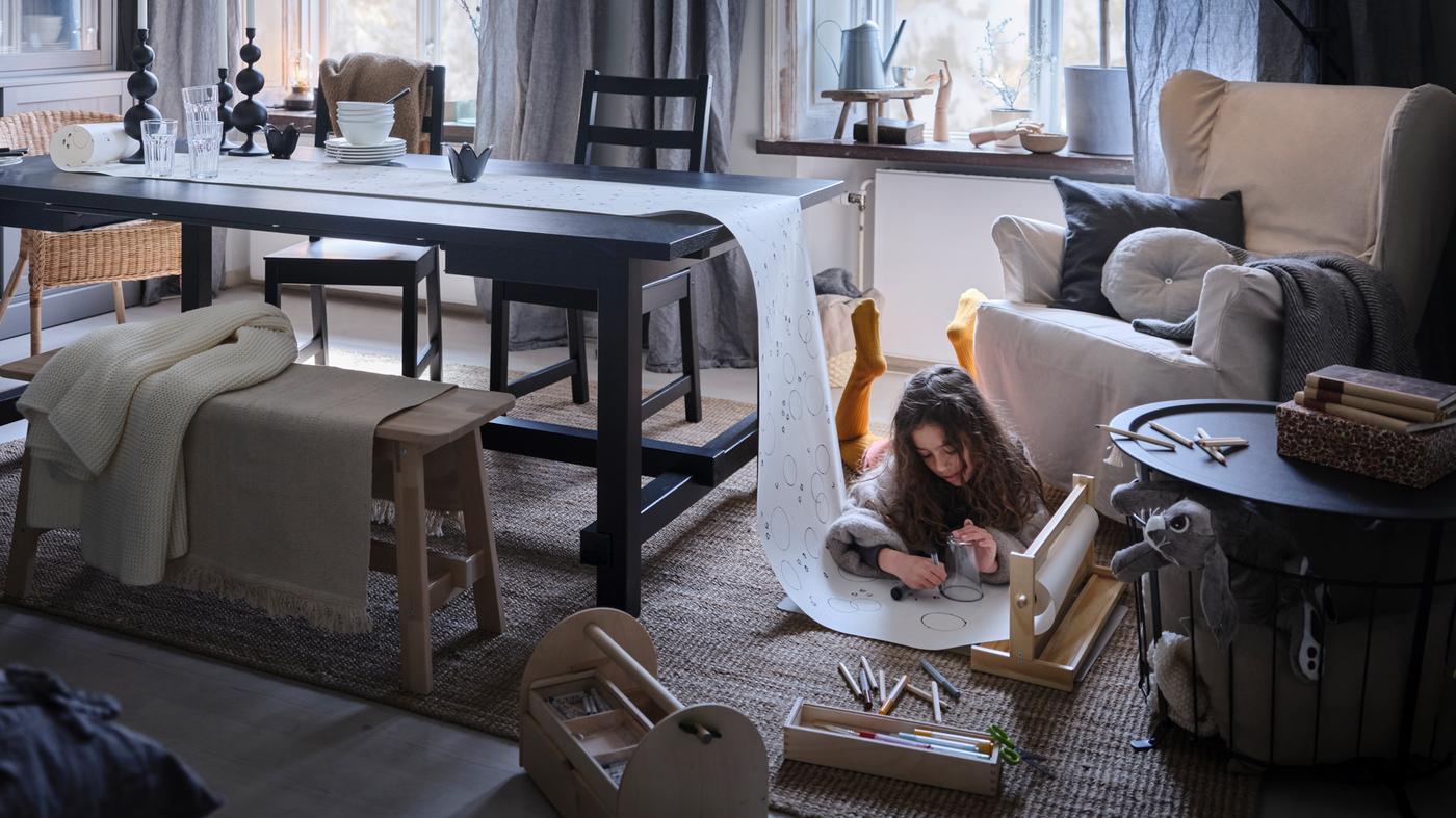 Una niña en el suelo con una alfombra dibujando en un rollo de papel, con un extremo sobre una mesa extensible negra NORDVIKEN .